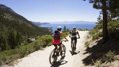 Two cyclists ride on a dirt path overlooking a lake surrounded by mountains and trees under a clear blue sky.