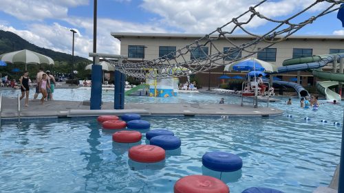 A pool area with colorful floating obstacles, water slides, and people enjoying the water under a cloudy sky.