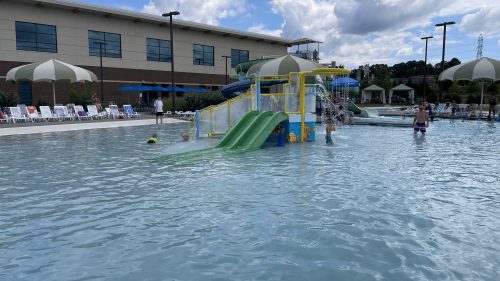 A water park scene with a green slide, children playing in the pool, and lounge chairs under umbrellas in the background.