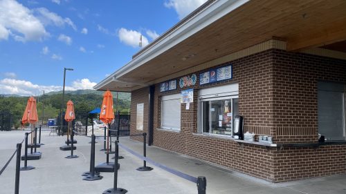 A concession stand with orange umbrellas and a queue area, set against a backdrop of blue skies and mountains.