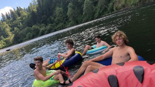 Four young people floating on colorful inner tubes in a river, surrounded by trees and greenery.