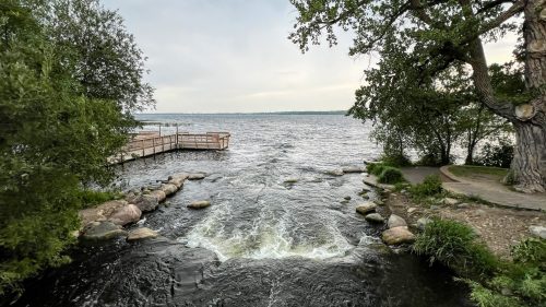 A serene view of a river flowing into a lake, surrounded by trees and a wooden dock in the distance.
