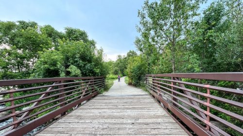 A wooden bridge leads into a lush green path, with a person walking in the distance.