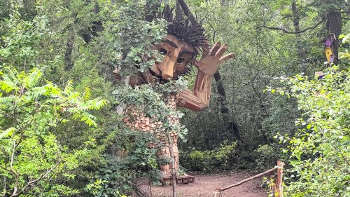 A large wooden figure with a friendly expression stands among trees, waving in a lush green forest.