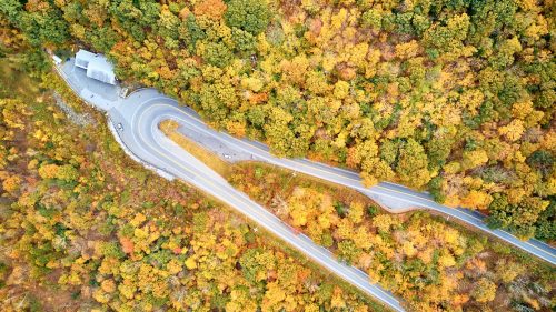 Aerial view of winding roads through vibrant autumn foliage with a small building nestled among the trees.