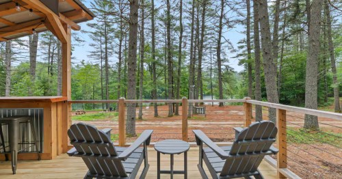 Two chairs with a table between them sit on the porch of the woodsy Pond+Pine glamping cabin in Oxford, Maine.