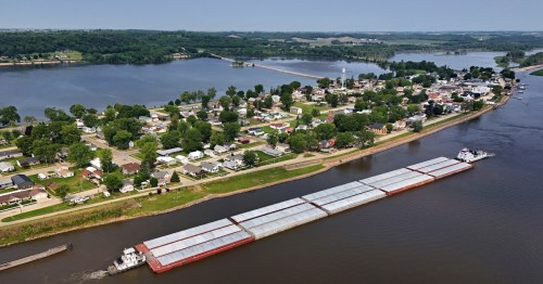 A barge floats past an island filled with homes and green trees on a river