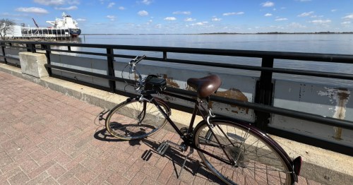A bicycle parked by a metal railing overlooks a wide river with a large cargo ship in the distance.