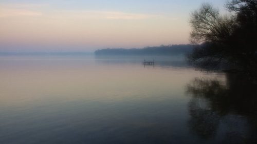 A serene lake at dawn, with soft pastel colors and mist rising from the water, reflecting trees along the shore.