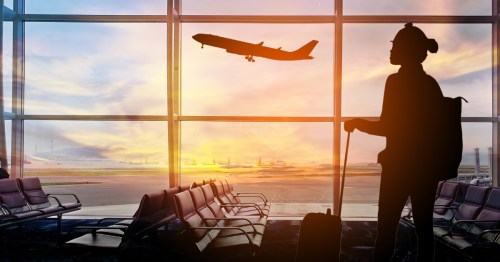 The silhouette of a person standing at the airport with luggage as a plane takes off in the distance.