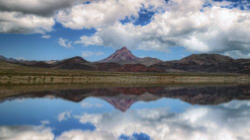 A serene landscape featuring a mountain reflected in calm water, under a blue sky with fluffy white clouds.