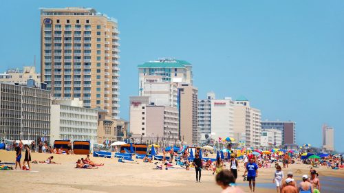 Crowded beach scene with people, colorful umbrellas, and tall buildings in the background under a clear blue sky.