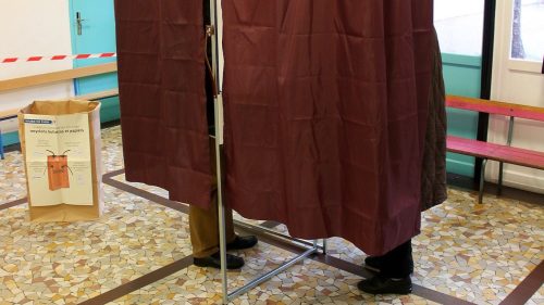 A voting booth with maroon curtains, partially obscuring two individuals in a brightly lit room with tiled flooring.