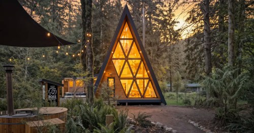 An A-frame cabin Airbnb in Oregon, with a wall of windows, is brightly lit up at dusk. The rental is surrounded by a lush forest.