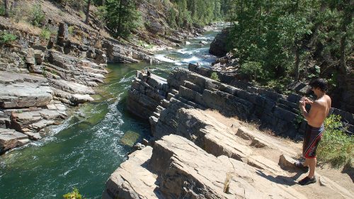A shirtless person stands on rocky terrain by a river, surrounded by trees and steep cliffs. Water flows nearby.