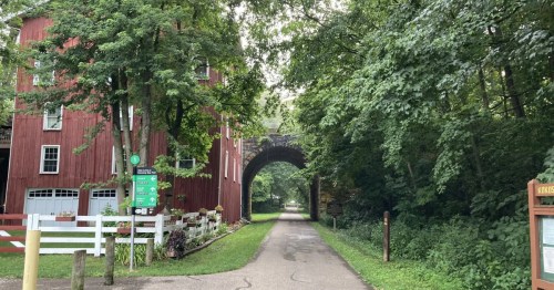 Tree-lined bike trail in Ohio passing a red historic building and stone railroad arch.