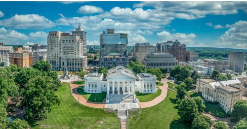 Aerial view of Downtown Richmond, Virginia, with the Virginia Capitol Building in the middle surrounded by green lawn and skyline under blue sky.