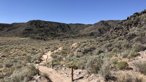A winding dirt path through a grassy landscape, surrounded by hills under a clear blue sky.
