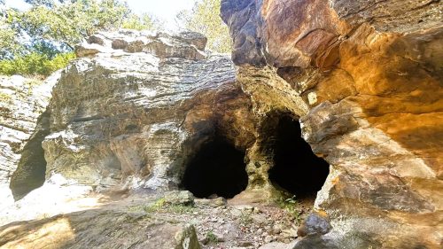 A rocky cave entrance surrounded by natural stone formations and greenery, with dark openings leading into the cave.
