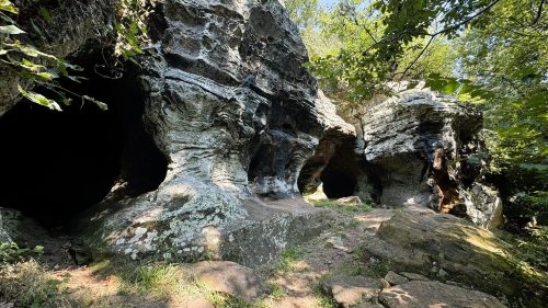 Rocky cave formations surrounded by greenery and trees under a clear blue sky.
