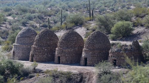 Four stone kilns with dome-shaped roofs, set against a desert landscape with cacti and shrubs.