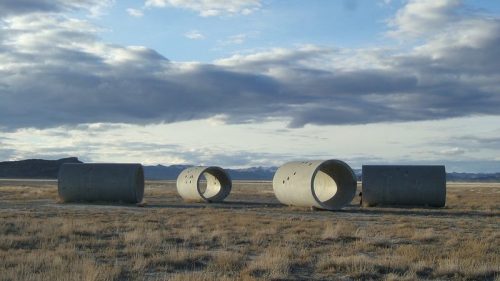 Four large concrete cylinders lie on a barren landscape under a cloudy sky, with distant mountains in the background.