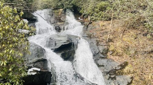 A serene waterfall cascades over rocky steps, surrounded by lush greenery and trees in a tranquil forest setting.