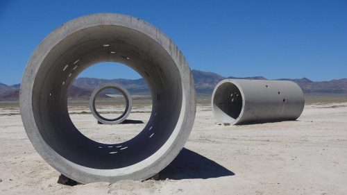 Three large concrete pipes stand on a dry, barren landscape under a clear blue sky, with mountains in the background.