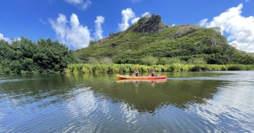 Kayakers paddling across the calm Wailua River in Hawaii, with lush hillside backdrop.