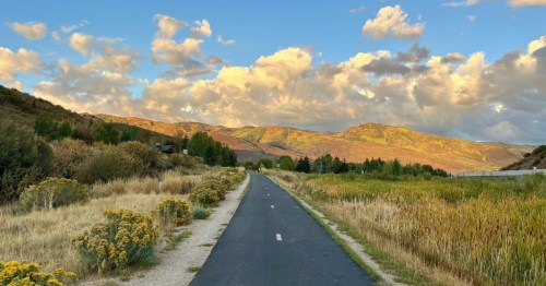 A paved rail trail in Utah going through open prairie with mountains and a big sky overhead.