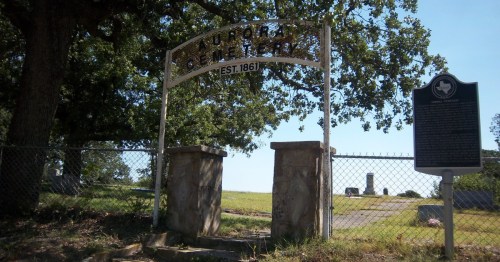 Entrance sign to Aurora Cemetery in Aurora, Texas, framed by trees and historic stone pillars.