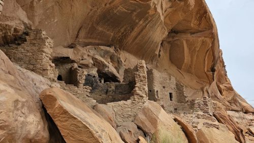 Ancient stone ruins nestled under a large rock overhang, surrounded by rugged terrain and sparse vegetation.