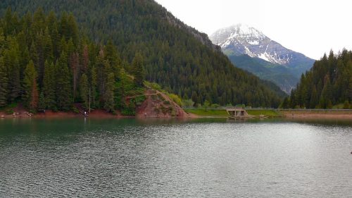 A serene lake surrounded by lush green mountains, with a snow-capped peak in the background under a cloudy sky.