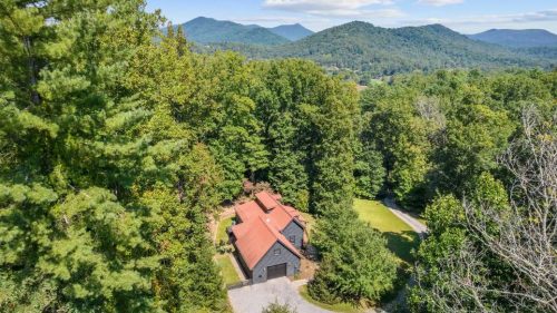 Aerial view of a house surrounded by lush trees and mountains under a clear blue sky.