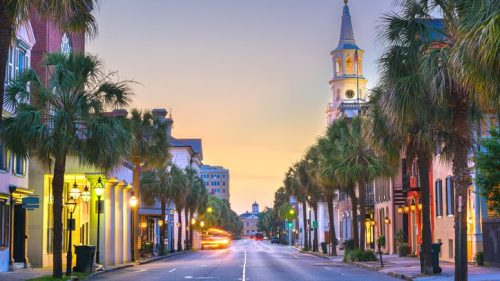 A picturesque street lined with palm trees, featuring historic buildings and a church steeple at sunset.