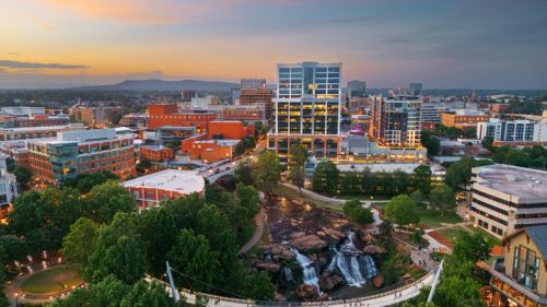 Aerial view of a city skyline at sunset, featuring a river, waterfall, and a pedestrian bridge surrounded by greenery.