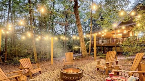 Cozy outdoor space with string lights, a fire pit, and wooden chairs surrounded by trees and a cabin in the background.