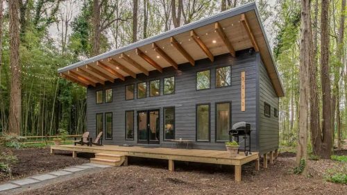 Modern black house with large windows, wooden deck, and outdoor seating, surrounded by trees and gravel pathway.