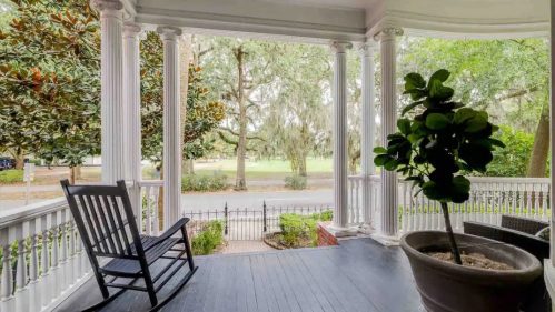 A porch with white columns, a rocking chair, and a potted plant, overlooking a tree-lined street.