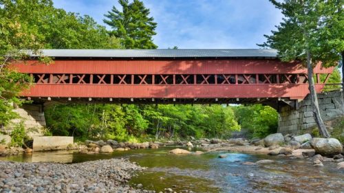 A red covered bridge spans a river, surrounded by lush green trees and rocky riverbanks under a clear blue sky.