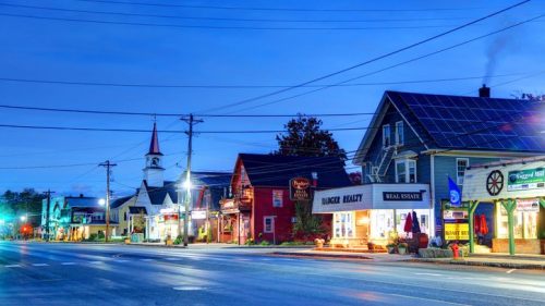 A quiet street at dusk featuring shops, a church with a steeple, and solar panels on a nearby building.