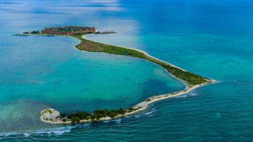 Aerial view of a narrow, winding island surrounded by turquoise waters, with a historic building in the background.
