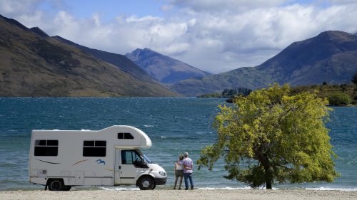 A couple stands by a lake, with mountains in the background and a camper van parked nearby under a tree.