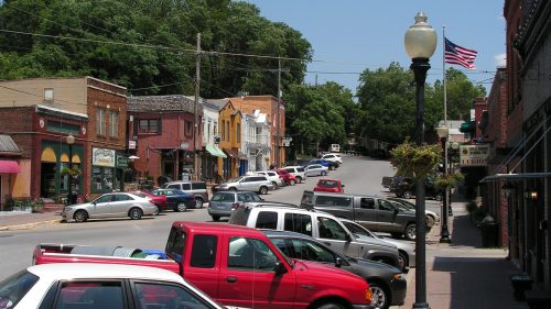 A quaint town street lined with shops and parked cars, featuring greenery and an American flag in the background.