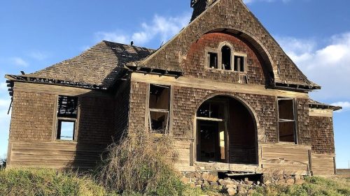 An abandoned, weathered wooden building with a steeple, surrounded by overgrown grass under a blue sky.