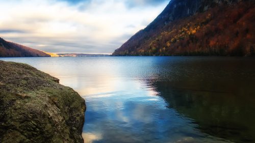A serene lake surrounded by mountains, reflecting colorful autumn foliage under a cloudy sky.