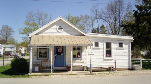 A small white house with a blue door and a metal roof, surrounded by trees and a gravel path.