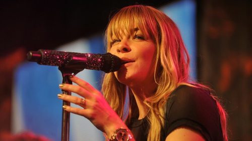 A singer with long blonde hair performs on stage, holding a sparkling microphone and singing passionately.