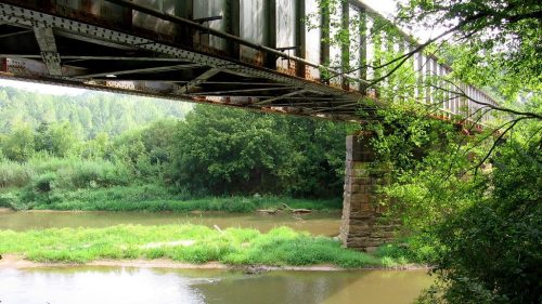 A rusted train bridge spans a calm river, surrounded by lush greenery and trees on both banks.