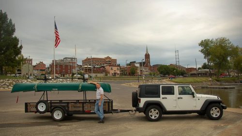 A man pulls a trailer with a canoe behind a white Jeep at a waterfront, with a small town and cloudy sky in the background.